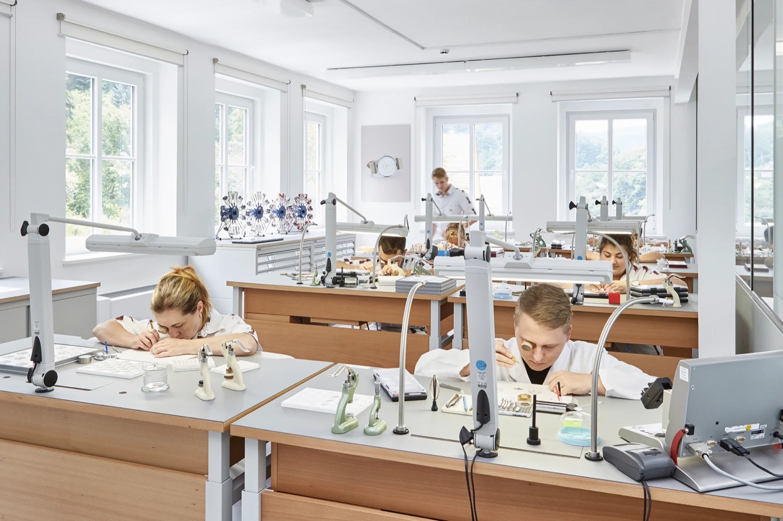 A watchmaker at a bench in Glashütte assembling a movement with tweezers under bright light, wearing a loupe